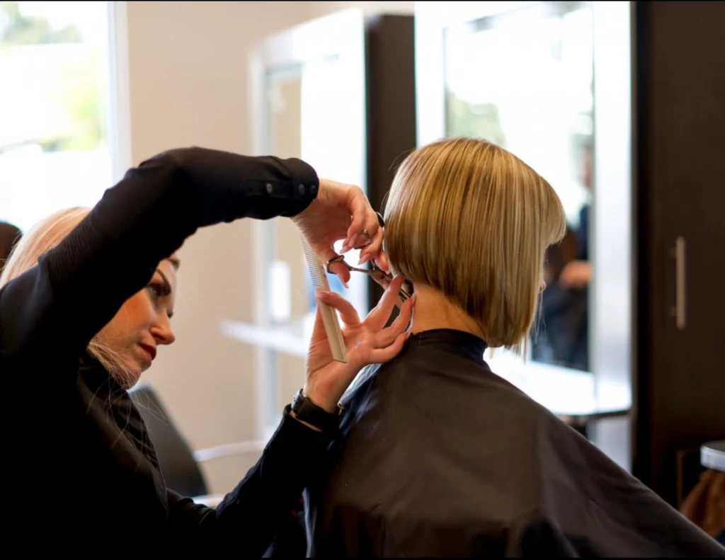 A hairstylist is cutting the back of a client's short, blonde hair in a salon. The client is wearing a black cape. - Gila Rut Salon | San Diego and Chula Visa, CA