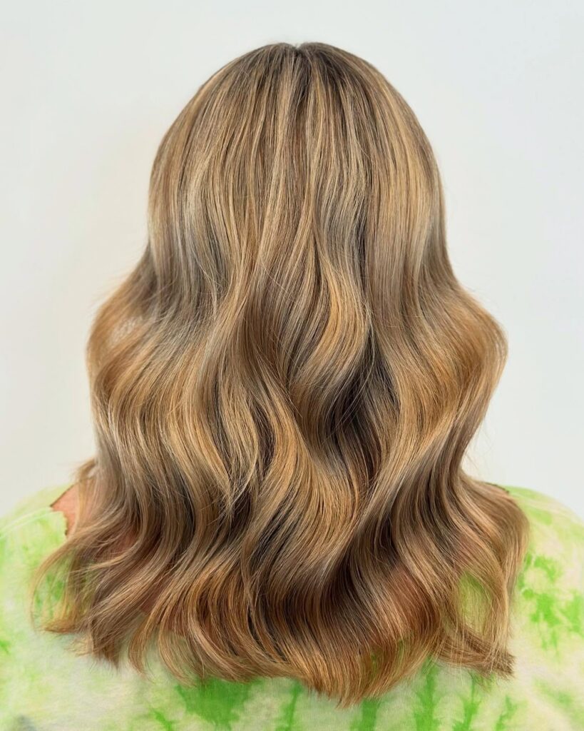 A person with long, wavy, blonde hair seen from behind, wearing a green tie-dye shirt against a plain light background. - Gila Rut Salon | San Diego and Chula Visa, CA