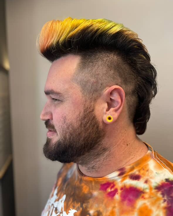 A man with a thick beard and colorful mohawk hairstyle, featuring yellow, orange, and green hues, and wearing a tie-dye shirt and yellow plug earring. - Gila Rut Salon | San Diego and Chula Visa, CA