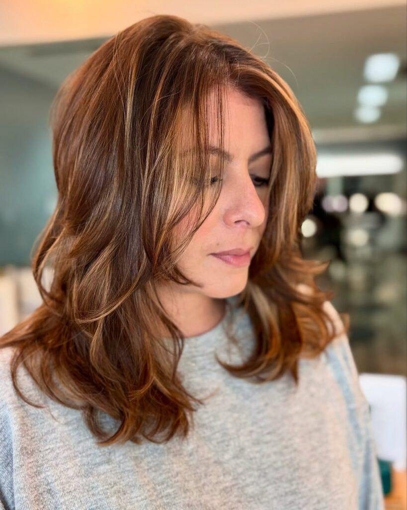 Woman with shoulder-length, layered brown hair and subtle highlights looking down, wearing a light gray top in an indoor setting. - Gila Rut Salon | San Diego and Chula Visa, CA