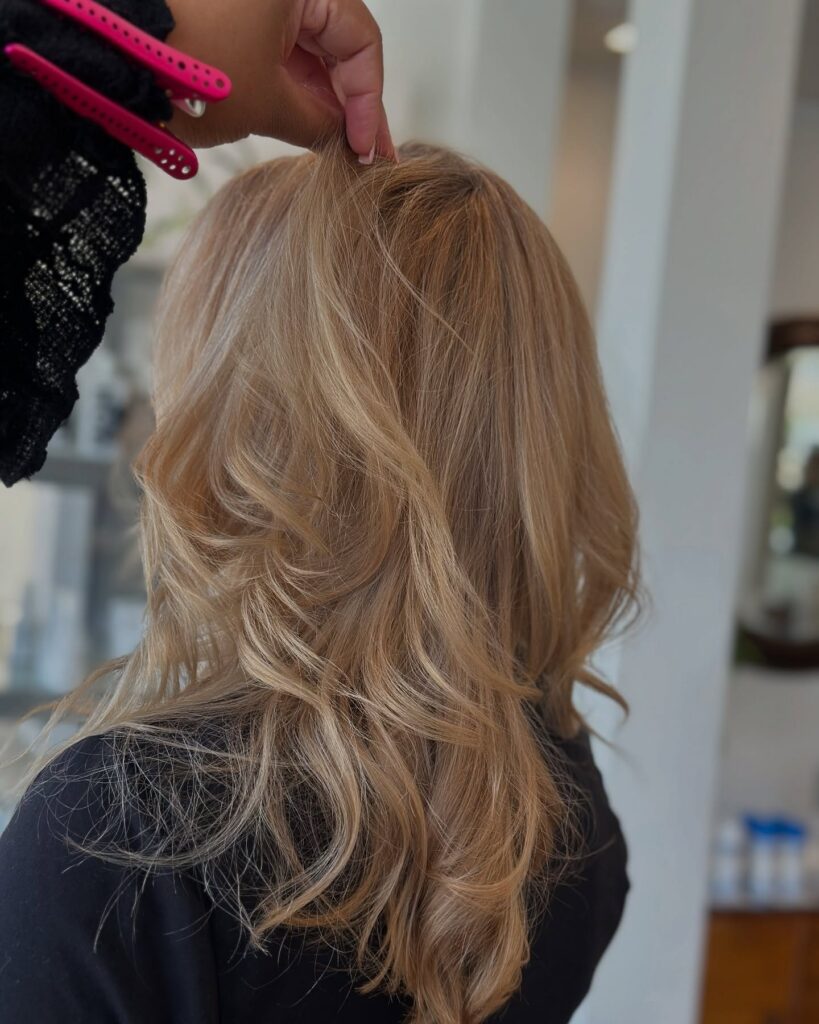 Person with long, wavy blonde hair is shown from the back in a salon as another hand lifts a section of their hair. - Gila Rut Salon | San Diego and Chula Visa, CA