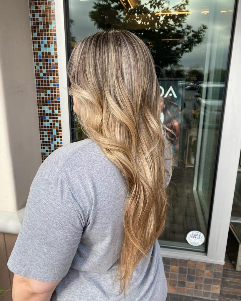 A person with long, wavy blonde hair and darker roots stands outside a building, facing away from the camera. - Gila Rut Salon | San Diego and Chula Visa, CA
