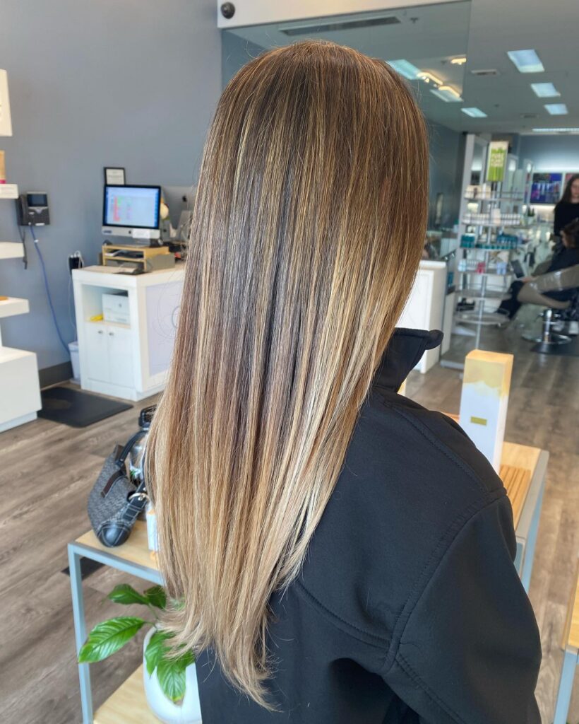 A person with long, straight, highlighted hair is standing in a modern salon, facing away from the camera. - Gila Rut Salon | San Diego and Chula Visa, CA