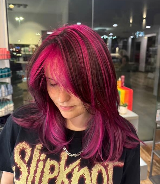 Person with straight, shoulder-length hair dyed dark brown with vibrant pink highlights, wearing a black Slipknot band t-shirt, standing in a well-lit indoor setting. - Gila Rut Salon | San Diego and Chula Visa, CA