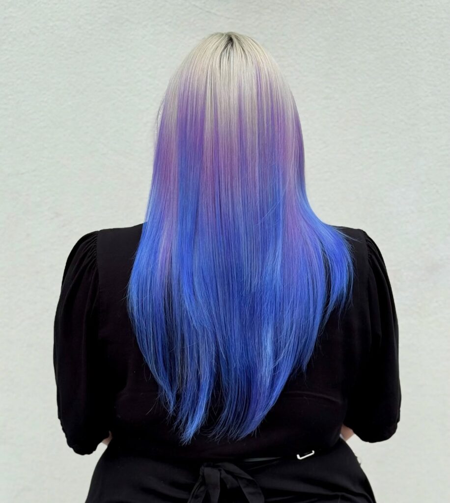 Person with long, straight hair dyed platinum blonde at the roots, fading into pastel purple and vibrant blue, shown from behind against a plain light background. - Gila Rut Salon | San Diego and Chula Visa, CA