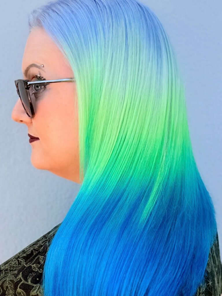 Person with long, straight hair dyed in a gradient of light blue at the roots, transitioning to bright green and deep blue at the ends, wearing glasses and dark lipstick. - Gila Rut Salon | San Diego and Chula Visa, CA