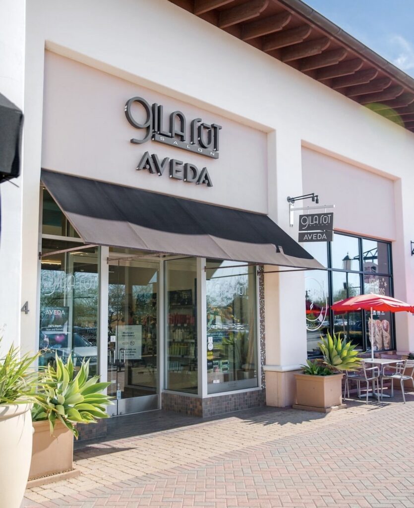 Exterior view of a Gila Rut Aveda salon with large windows, black awning, outdoor plants, and patio seating with red umbrellas. - Gila Rut Salon | San Diego and Chula Visa, CA