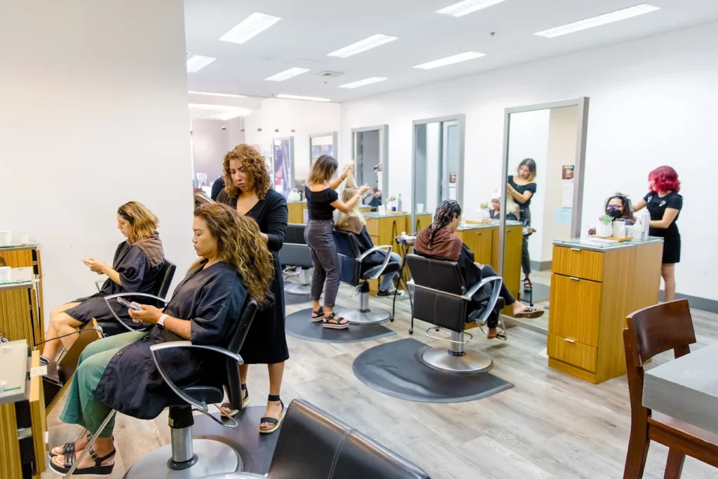 Several people are getting their hair styled in a modern salon. Stylists work at individual stations while clients sit in chairs, some using their phones. - Gila Rut Salon | San Diego and Chula Visa, CA