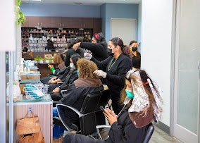 Several people wearing masks receive hair treatments from stylists in a modern salon; one client looks at her phone while others sit at stations. - Gila Rut Salon | San Diego and Chula Visa, CA