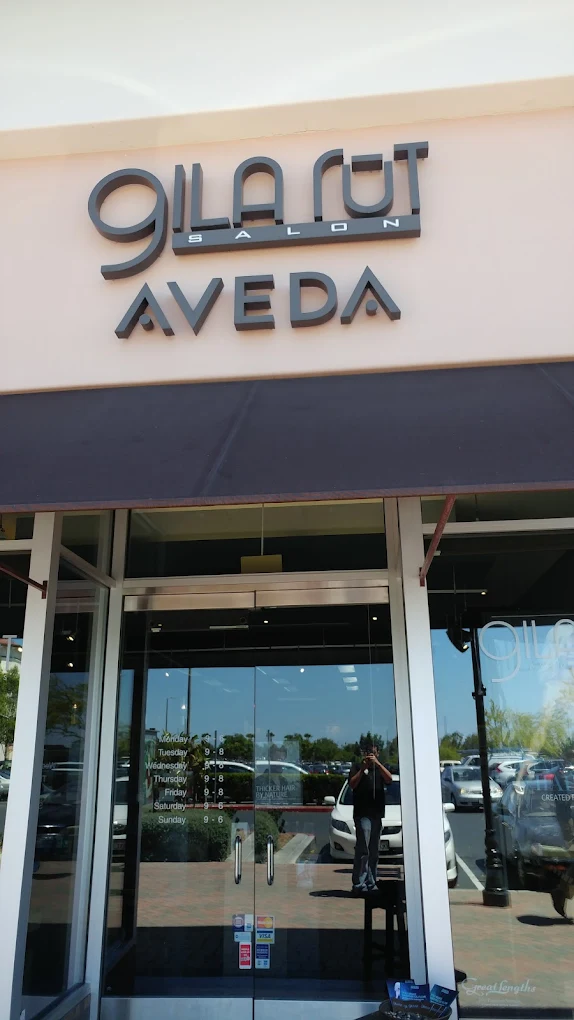Storefront of Gila Rut Salon Aveda with glass doors, black awning, and visible reflections of parked cars and a person taking a photo. - Gila Rut Salon | San Diego and Chula Visa, CA